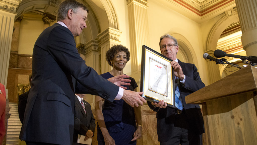 Bob Murphy, AARP Colorado state director (right to left), and Jean Nofles, AARP Colorado state president hands a framed recognition to Governor John Hickenlooper, Sept. 18, 2018. (Kevin J. Beaty/Denverite)