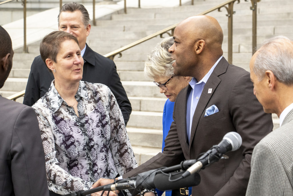 Mayor Michael Hancock and Piep van Heuven shake hands, with Councilwoman Mary Beth Susman in the background, after a press conference to announce the creation of Denver's Department of Transportation and Infrastructure on the City and County Building steps, April 2, 2019. (Kevin J. Beaty/Denverite)