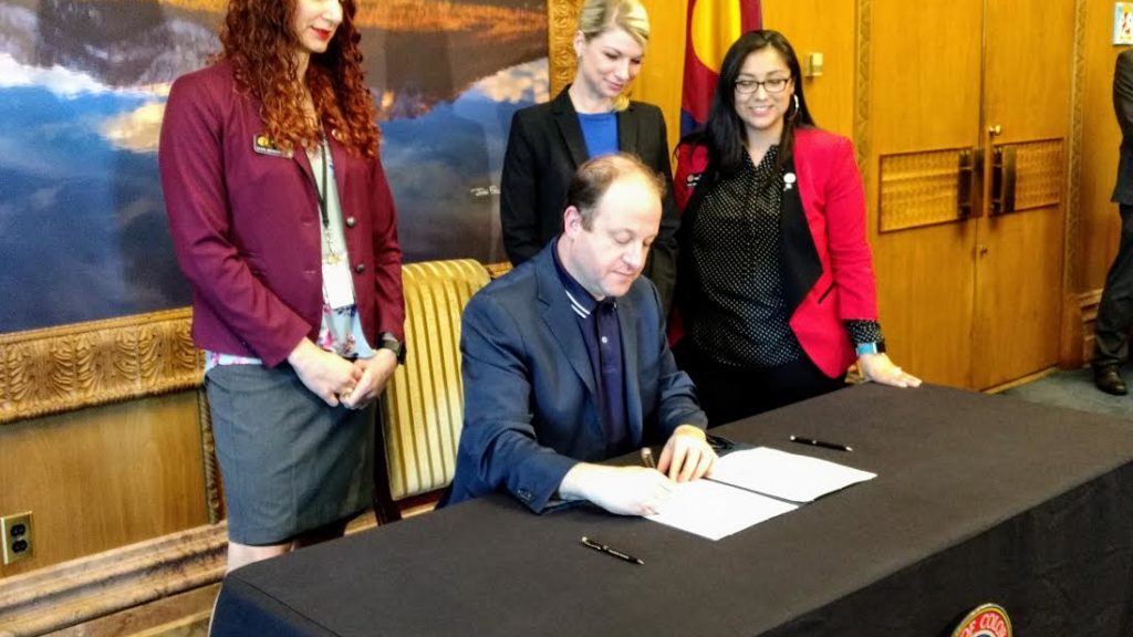Democratic representatives Brianna Titone and Brittany Petterson of Jefferson County and Serena Gonzales-Gutierrez of Denver, standing from left to right look on as Gov. Jared Polis signs the law they sponsored on rental applications on April 25, 2019. (Donna Bryson/Denverite)