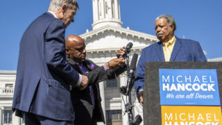 Former Governor John Hickenlooper and Mayor Michael Hancock try to fix a microphone for former Mayor Wellington Webb during a rally for Hancock's reelection campaign, May 14, 2019. (Kevin J. Beaty/Denverite)