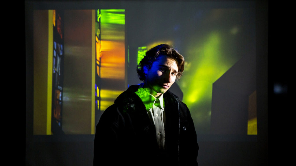 Myles Goldstein, Denver Jewish Film Festival entrant, poses for a portrait beneath a frame from his documentary inside a University of Denver media classroom. Jan. 28, 2020. (Kevin J. Beaty/Denverite)