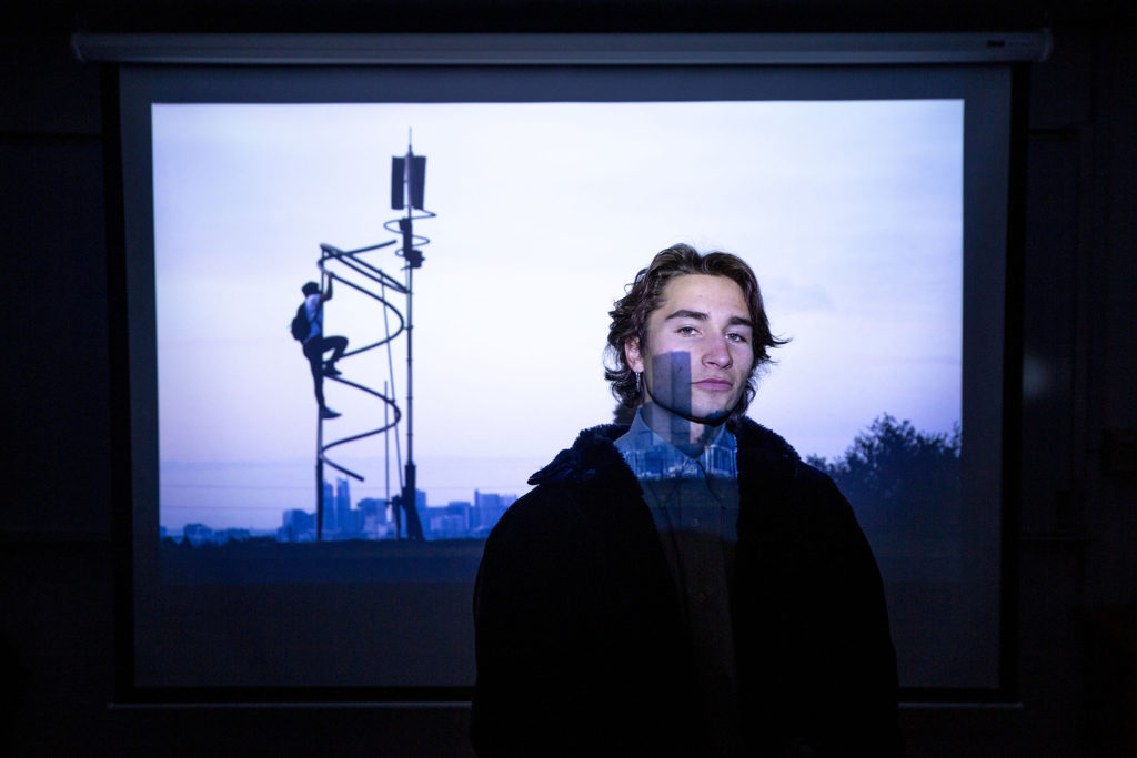 Myles Goldstein, Denver Jewish Film Festival entrant, poses for a portrait beneath a frame from his documentary inside a University of Denver media classroom. Jan. 28, 2020. (Kevin J. Beaty/Denverite)