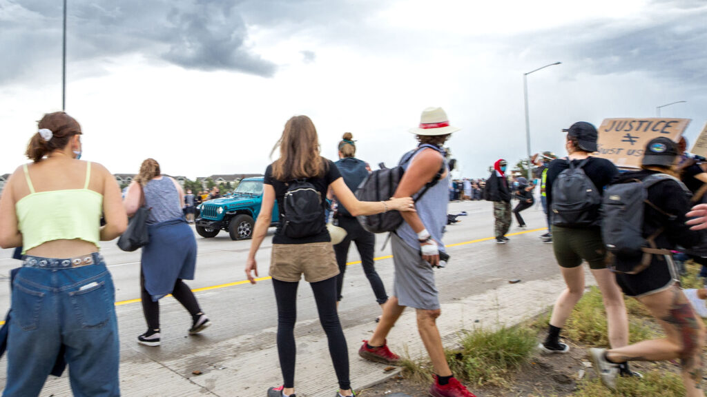 A driver in a blue Jeep speeds through a protest demanding justice for Elijah McClain. A protester fired a gun as the vehicle drove by, shooting other protesters on July 25, 2020.