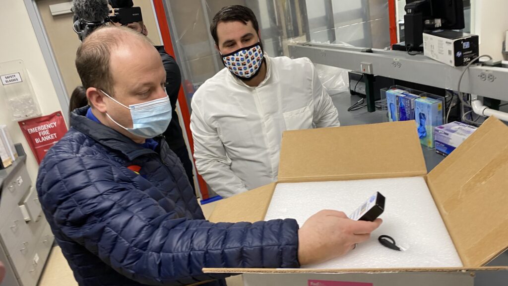 Gov. Jared Polis stands next to a box containing a COVID-19 vaccine on Monday, Dec. 14, at a state laboratory in Denver. (John Daley/CPR News)