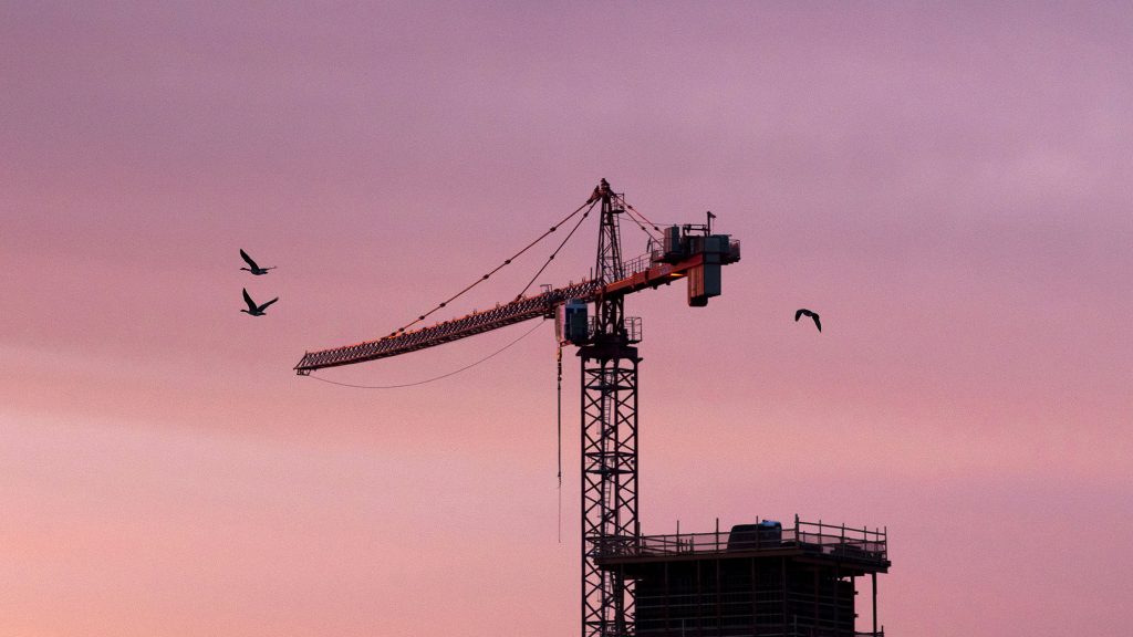 Canada Geese fly by cranes looming above Globeville Landing Park at dawn. April 23, 2021.