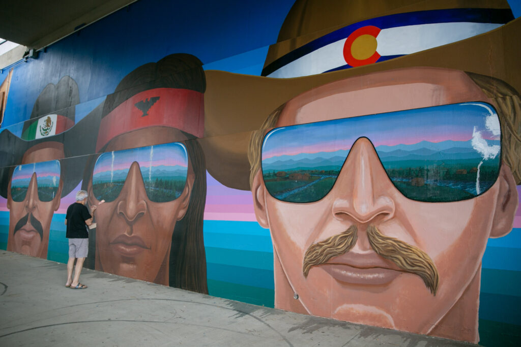 Emanuel Martinezstands in front of three large, painted faces on the wall, holding a paint brush. The dark-skinned figure on the far left wears a cowboy hat with a Mexican flag hatband, the dark-skinned figure in the middle wears a red headband with a thunderbird, and the light-skinned figure on the right wears a cowboy hat with a Colorado flag hatband.
