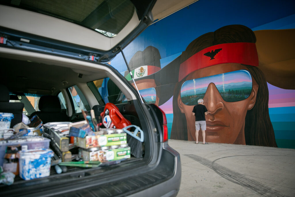 Emanuel Martinez uses light tan paint to touch up the nose of a large head painted on the wall. In the foreground, a hatchback Subaru is open, reveling a trrunk of paint supplies, jars, a broom head, a ladder, and more.