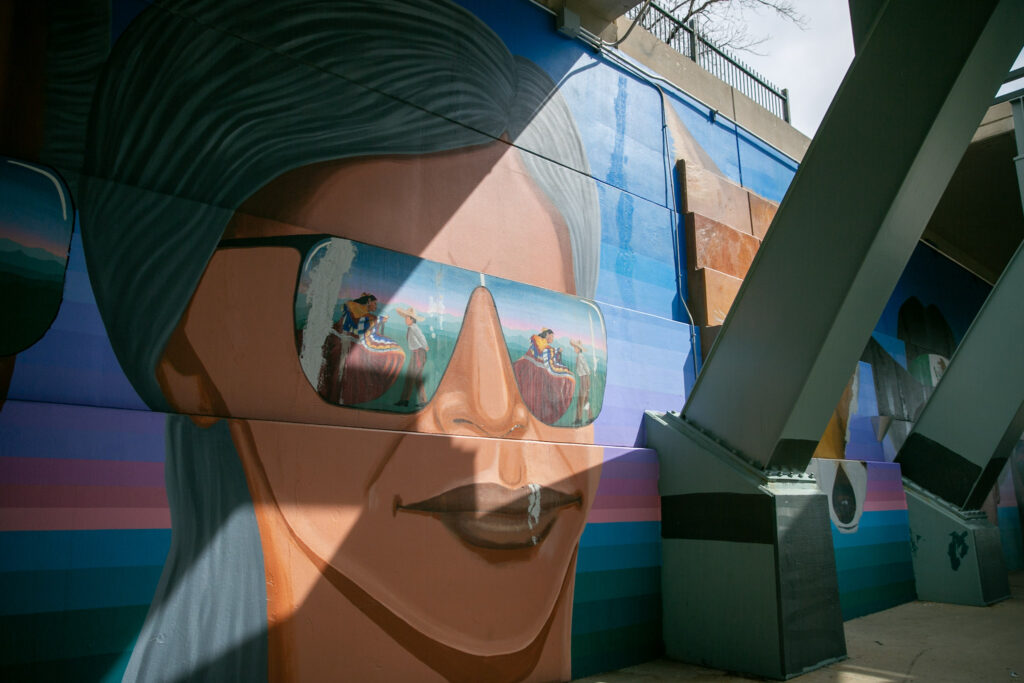 A sunglassed figure with dark skin and long black hair from Emanuel Martinez's "Confluent People." The person's lenses reflect a traditional Mexican ballet folklorico with a man and a woman standing in a field, preparing to dance.
