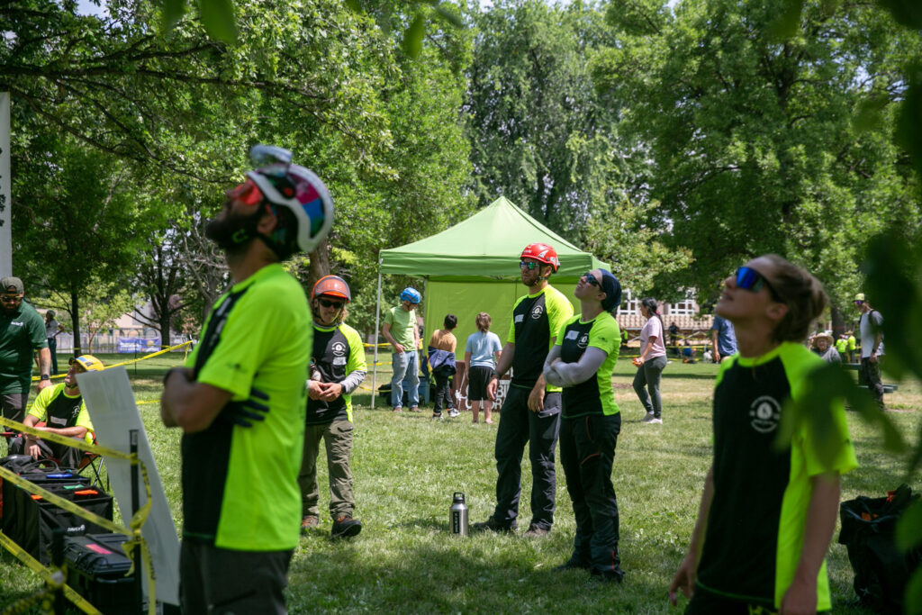 A crowd of competitors in vibrant yellow/green shirts look up at a tree, watching Wyatt Chipps (out of frame) competing in the throwline event.