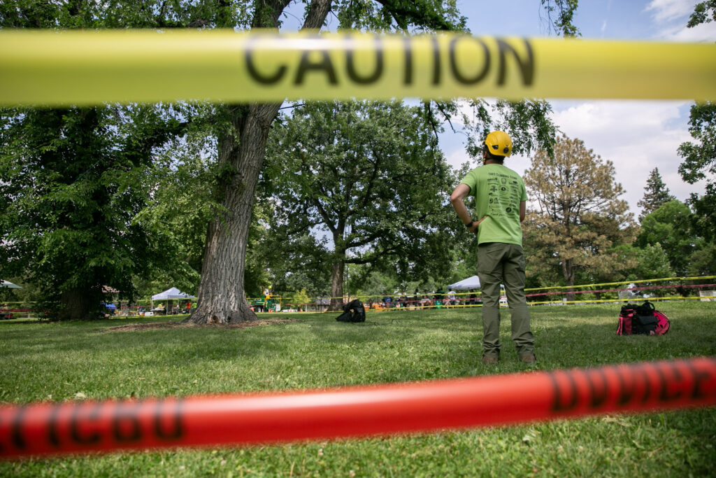 A man in a yellow helmet and green T-shirt looks up at a tree, framed by yellow and red caution tape.