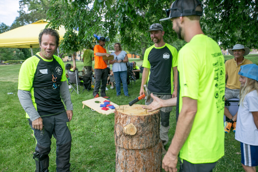 Patrick O'Meara stands in a circle around a stump with nails in it. He's playing hammerschlagen with two other men, one of whom is actively flipping the hammer over backwards before trying to hit one of the nails in the stump.