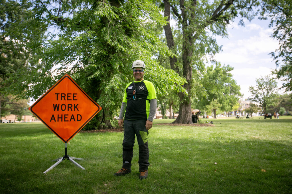 Patrck O'Meara wears a white helmet, vibrant shirt, and safety googles while posing next to an orange "TREE WORK AHEAD" sign in a park.