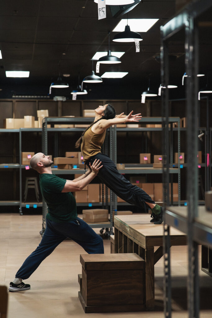 Two dancers are in a room full of cardboard boxes. A women falls forward from an elevated stage, arms stretched overhead, as a man standing on the ground catches her by the hips.