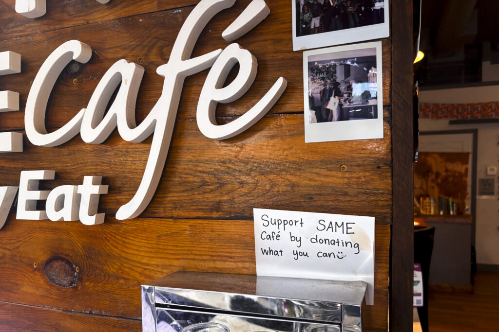 A sign above a donation box against a reclaimed wood wall that reads, "Support SAME cafe by donating what you can " with a smiley face.