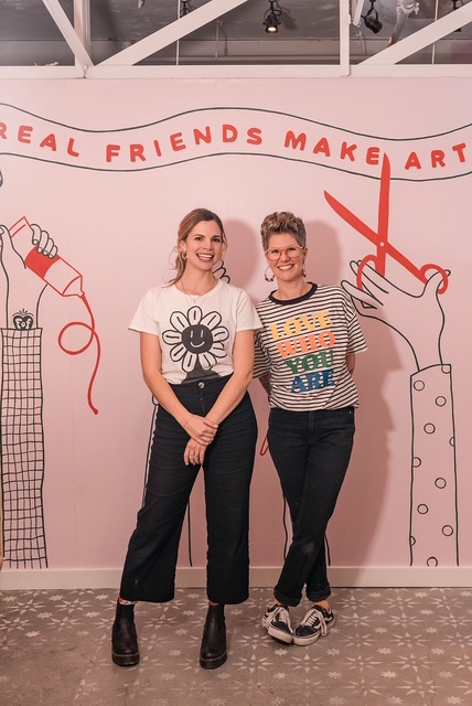 Two people wearing dark pants and graphic t-shirts stand in front of a pink wall. Painted on the wall are two illustrative arms holding art supplies and text reading, "Real Friends Make Art."